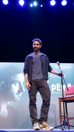 Humorista en escena con chaqueta azul, camiseta clara y vaqueros, cerca de un micrófono y un cartel con parte de su nombre y el título de su espectáculo.
