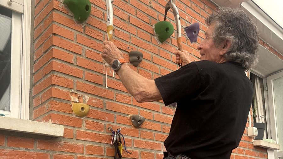 Alpinista mayor con pelo canoso escalando un rocódromo exterior con piolets y presas de colores;  a su alrededor se observan una ventana, una planta y material de escalada.
