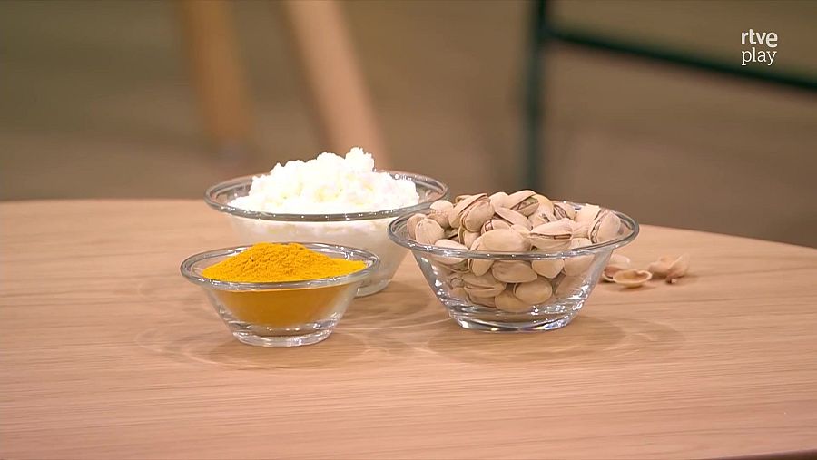 Cúrcuma en polvo, pistachos y requesón blanco en tres cuencos de cristal sobre una mesa de madera.  Predicción alimentaria para 2025.