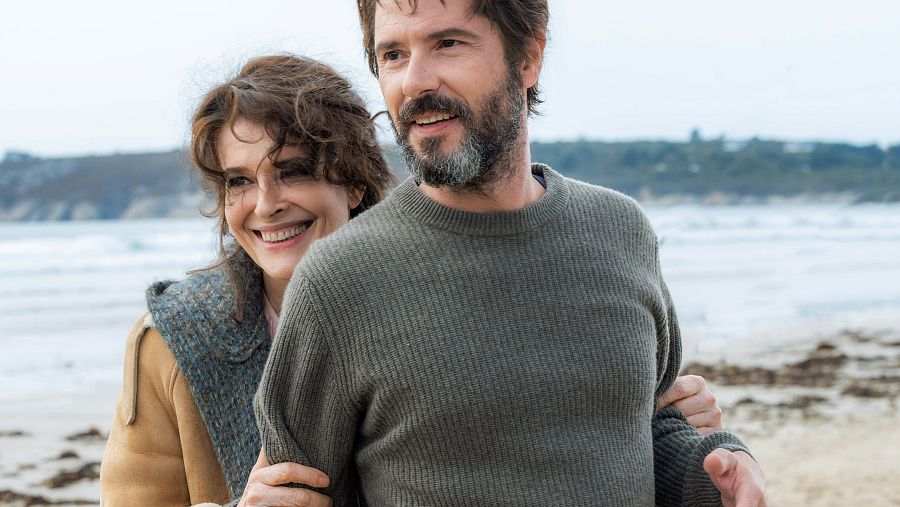 Pareja mayor abrazada en una playa, sonriendo a la cámara. Él viste jersey gris, ella chaqueta beige y bufanda.  Fondo marino y costa.