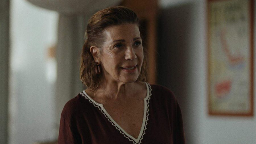 Actriz con blusa granate y pendientes, expresión serena, en interior con cuadro abstracto de fondo.