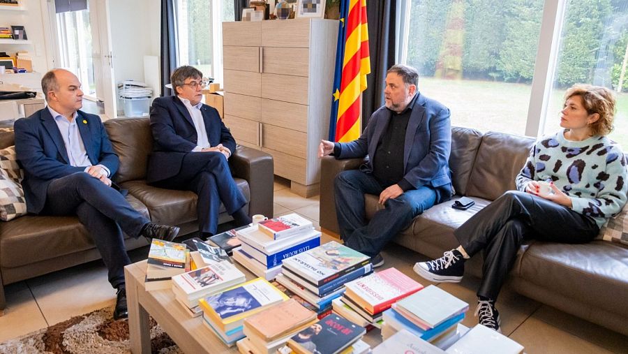 Cuatro personas, vestidas de manera informal y formal, reunidas en una sala con una bandera catalana al fondo.  Se observa una mesa con libros.