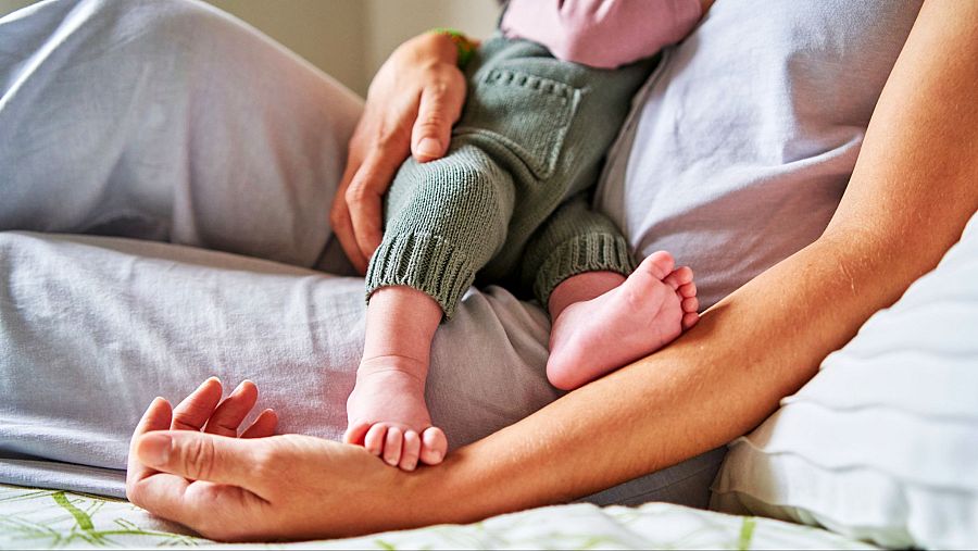 Madre e hijo comparten un momento íntimo y tranquilo. El bebé, con un pelele verde oliva, descansa en el regazo de su madre en una cama con sábanas de estampado vegetal.