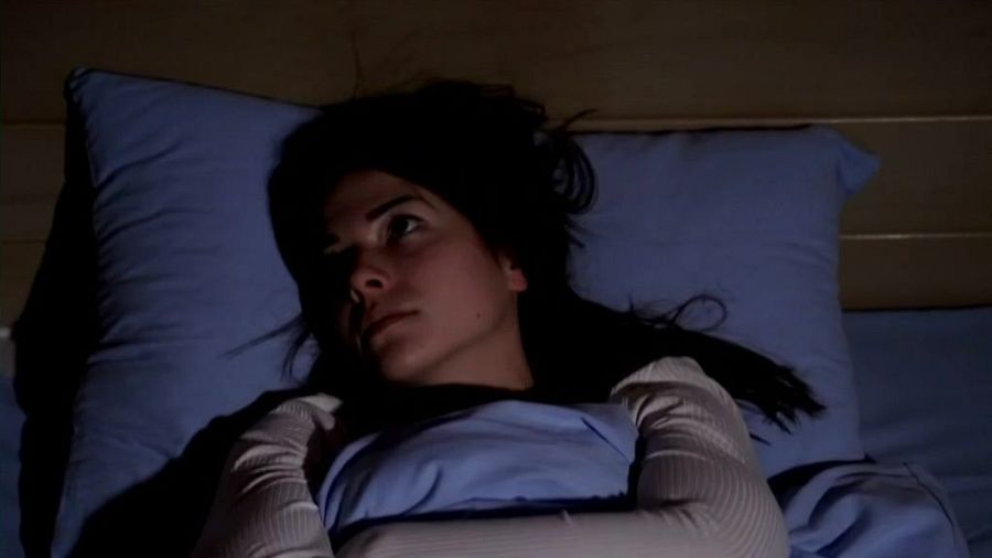 Mujer joven despierta en cama oscura, expresión preocupada.  Insomnio sugerido. Contexto: falta de sueño y enfermedades.