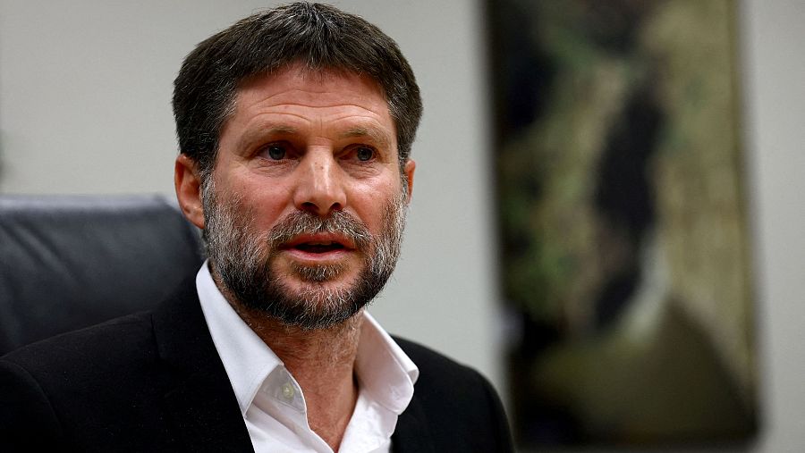 Imagen de Bezalel Smotrich, con camisa blanca y chaqueta oscura, expresión seria, posiblemente hablando, con una pintura borrosa de fondo.