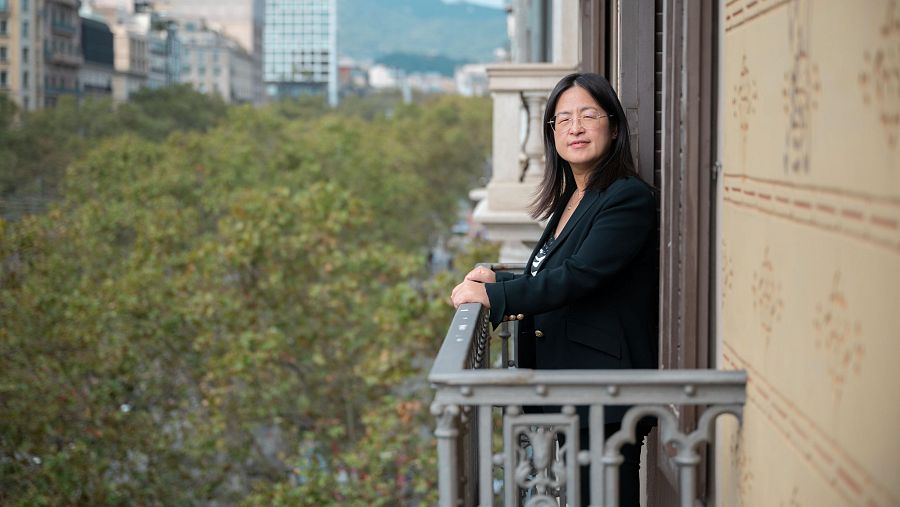 Mujer con gafas y chaqueta oscura en balcón de Barcelona, mirando hacia la izquierda; vista urbana al fondo.