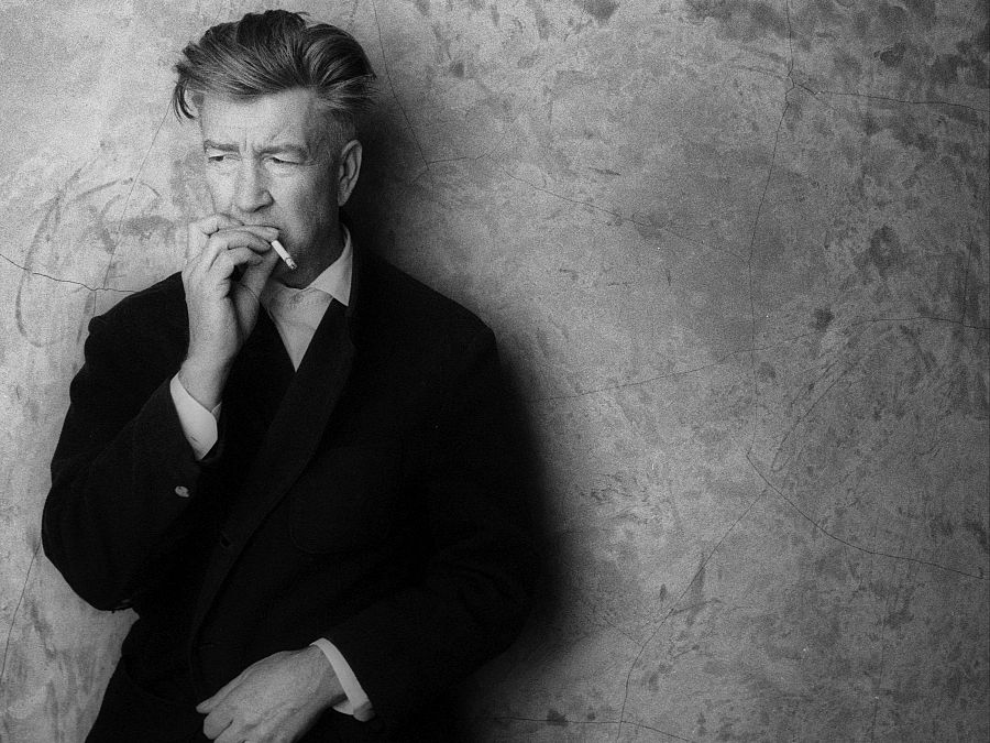 David Lynch, en blanco y negro, fuma un cigarrillo en su estudio. Traje oscuro, camisa blanca, expresión seria.  Foto de 2002.