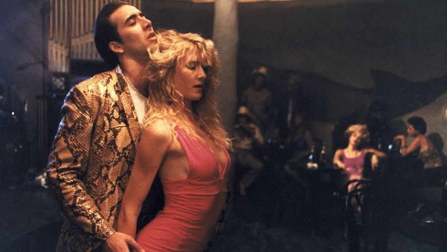 Nicolas Cage y Laura Dern bailando íntimamente en un bar. Él viste una chaqueta estampada y ella un vestido rosa corto.  Escena de película.