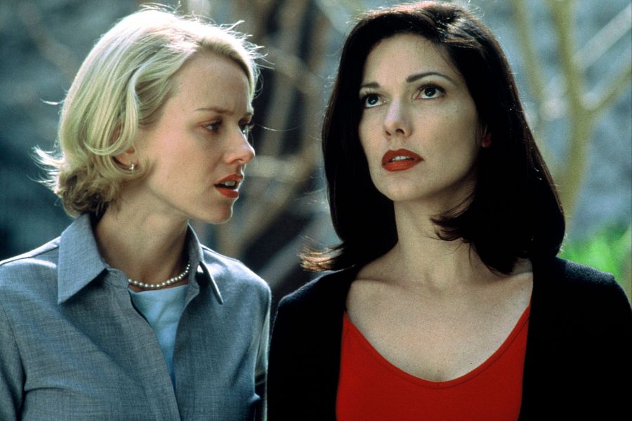 Escena de *Mulholland Drive*: Naomi Watts, con camisa gris y collar, y Laura Harring, con camiseta roja y chaqueta negra, interactúan con expresiones serias.