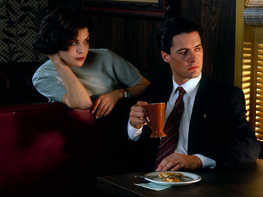 Sherilyn Fenn, con suéter claro, y Kyle MacLachlan, con traje oscuro, comparten una escena tranquila en un restaurante.  En la mesa hay café y pasteles.