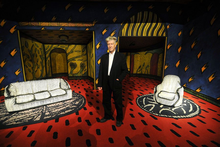 David Lynch, en traje negro, posa en una habitación con decoración surrealista: paredes azul y amarillo, suelo rojo, y muebles blancos de diseño estilizado.