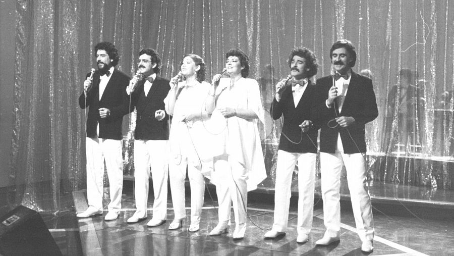 Grupo musical de seis personas (cuatro hombres y dos mujeres) en escena, con micrófonos, vestidos de oscuro y blanco. Imagen en blanco y negro, posiblemente de una actuación televisiva.