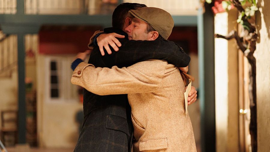 Emotiva despedida entre dos hombres: uno con boina y papel, el otro con chaqueta oscura. Fondo con puerta, ventana y planta trepadora.