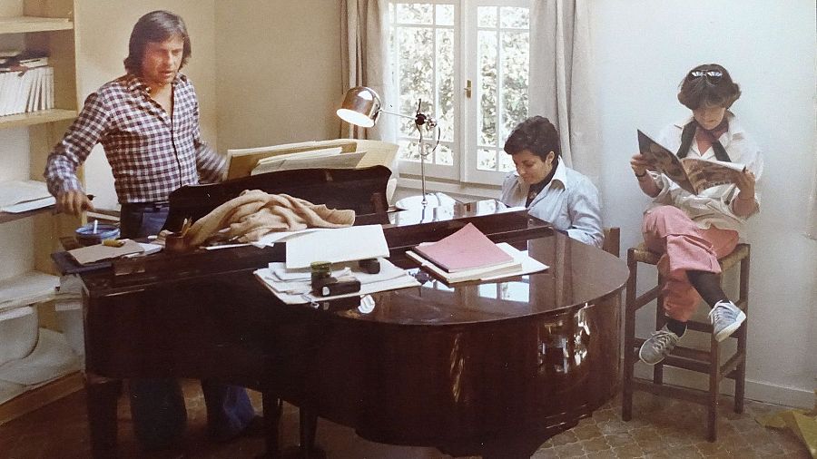 Miembros de Mocedades ensayan: un hombre dirige junto a un piano, una mujer toca y otra lee.  Partituras y una lámpara sobre el piano.