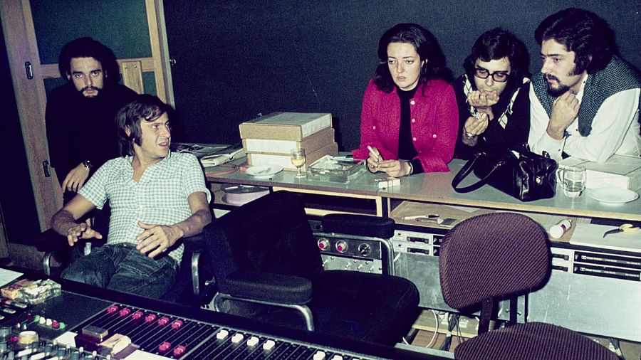 Estudio de grabación con seis personas; un hombre es entrevistado, otros observan.  Mesa de mezclas, equipo de producción vintage. Ambiente informal.