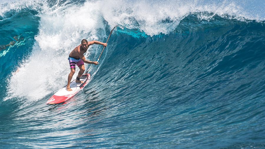 Hombre surfeando una ola grande en una tabla roja y blanca; viste pantalones cortos. Imagen con mensaje 