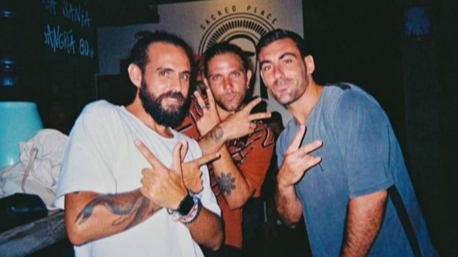 Tres hombres jóvenes, uno con barba y moño, posan haciendo el gesto de la paz.  La imagen, con poca luz, incluye el texto 