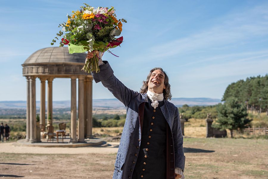 Actor con traje azul oscuro y ramo de flores, mostrando alegría.  Fondo con paisaje rural y arquitectura clásica. Despedida de rodaje.
