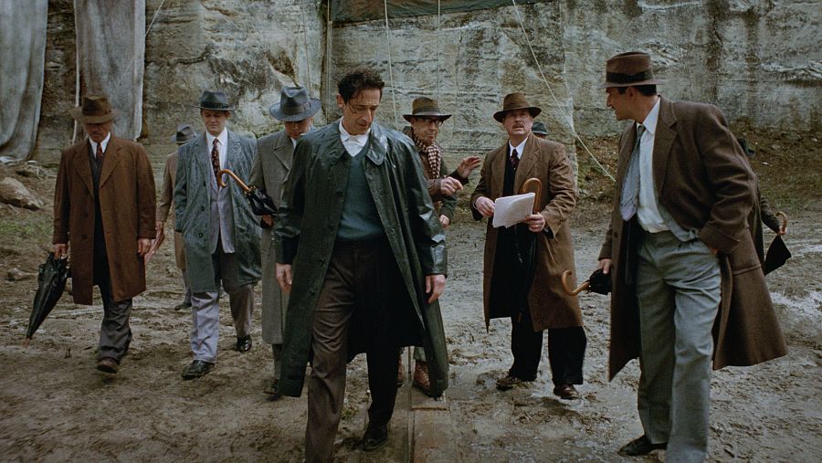 Ocho hombres con abrigos y sombreros caminan por un terreno embarrado, posiblemente en un entorno exterior en ruinas. Uno lleva un abrigo verde y papeles.