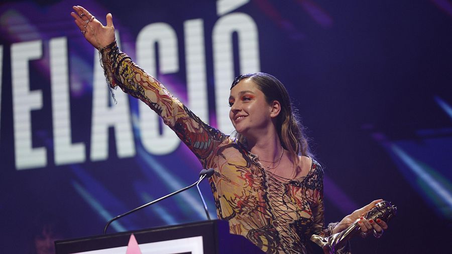 Actriz en escenario, sosteniendo un premio tras ganar el galardón a mejor interpretación revelación. Viste un vestido semitransparente multicolor y saluda al público.