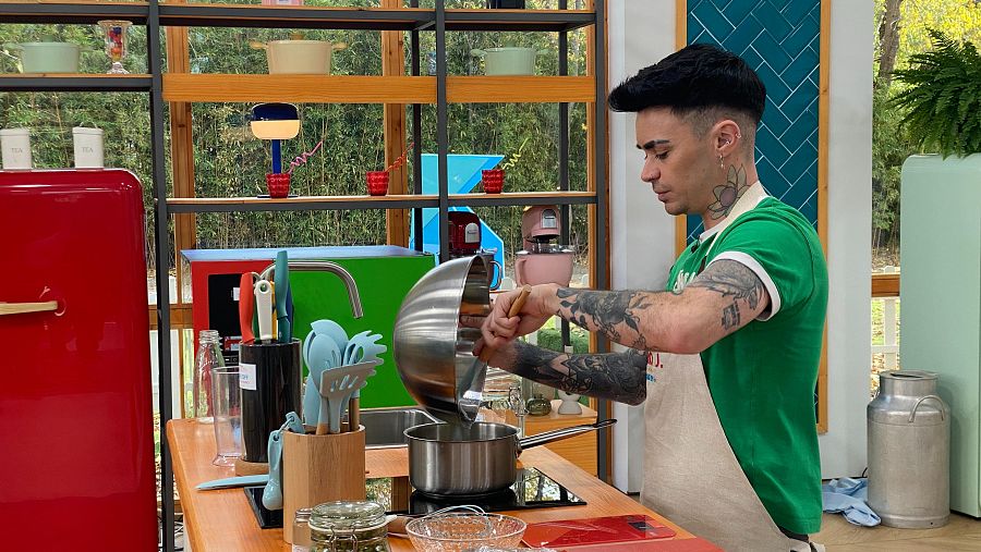 Mario Jefferson cocina baklava en un programa de repostería. Vierte líquido de un recipiente a una olla en una cocina colorida con una nevera roja y utensilios de cocina.