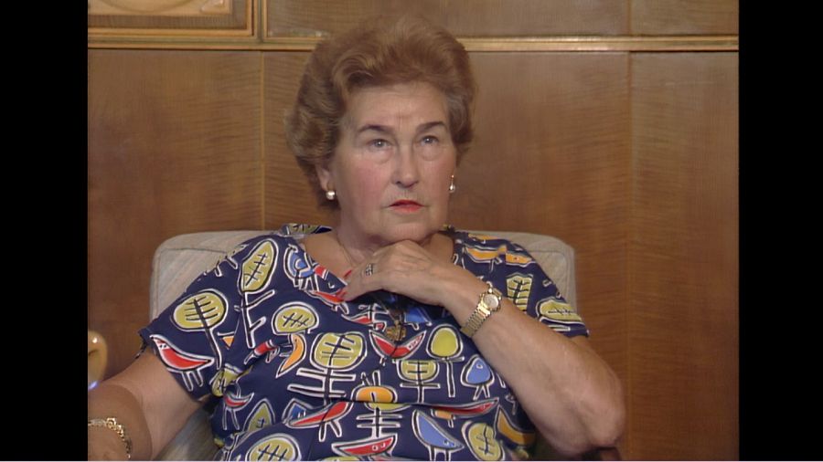 Mujer mayor con blusa estampada y joyas, sentada en sillón, expresión seria y pensativa.  Imagen en blanco y negro.