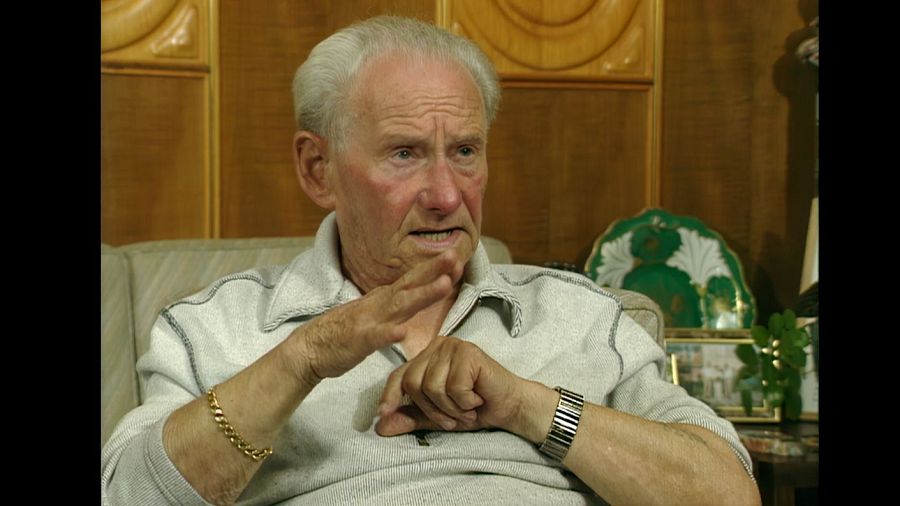 Hombre mayor, pelo canoso, camisa gris, gesticulando durante una entrevista en un ambiente doméstico.  Expresión seria.