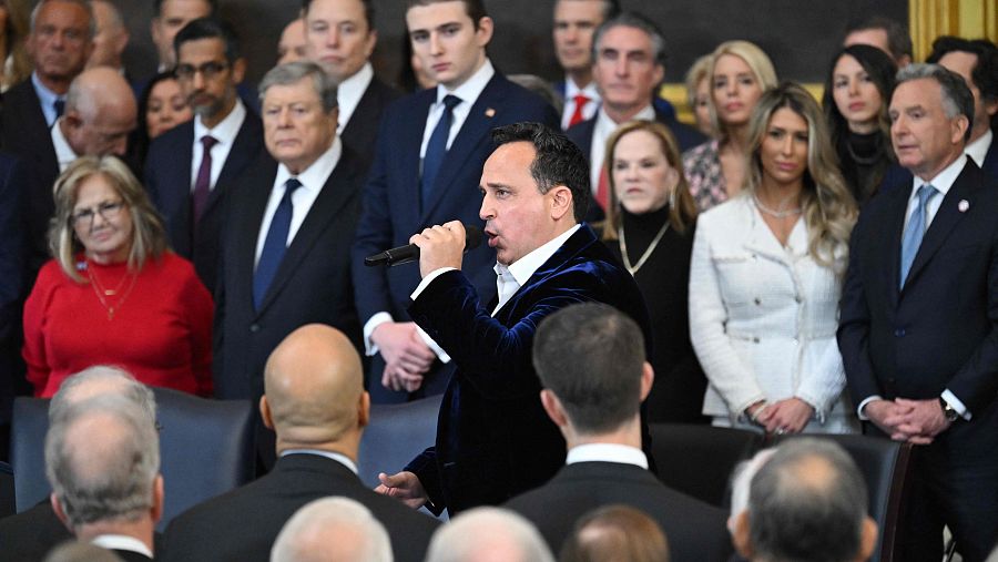 Cantante con chaqueta azul y micrófono actuando en una ceremonia en la Rotonda del Capitolio ante un público formalmente vestido.