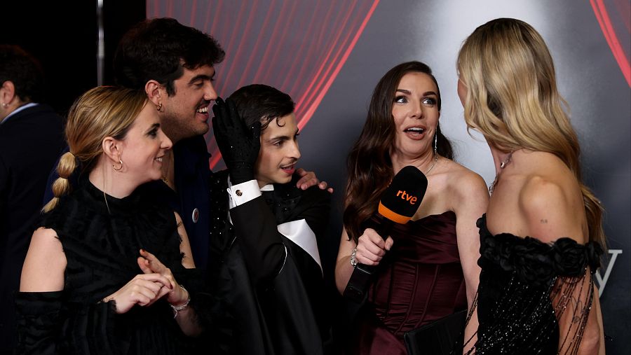 Inés Hernand entrevista en la alfombra roja de los Premios Goya 2024 a parte del equipo de 'Te estoy amando locamente'.  Ella viste un vestido granate y sostiene un micrófono.