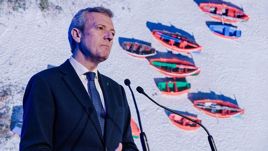 El presidente de Galicia, vestido de traje, habla ante micrófonos con un fondo de embarcaciones en una playa.  Posible entrevista o discurso.