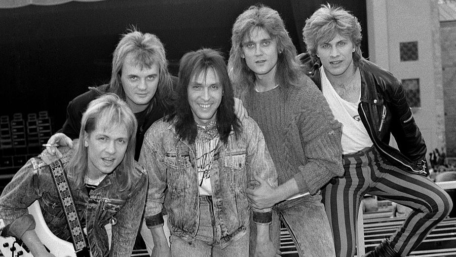 Cinco jóvenes con pelo largo y ropa de los 80 posan para una foto en blanco y negro; uno lleva una guitarra.  Posible foto promocional o de concierto.