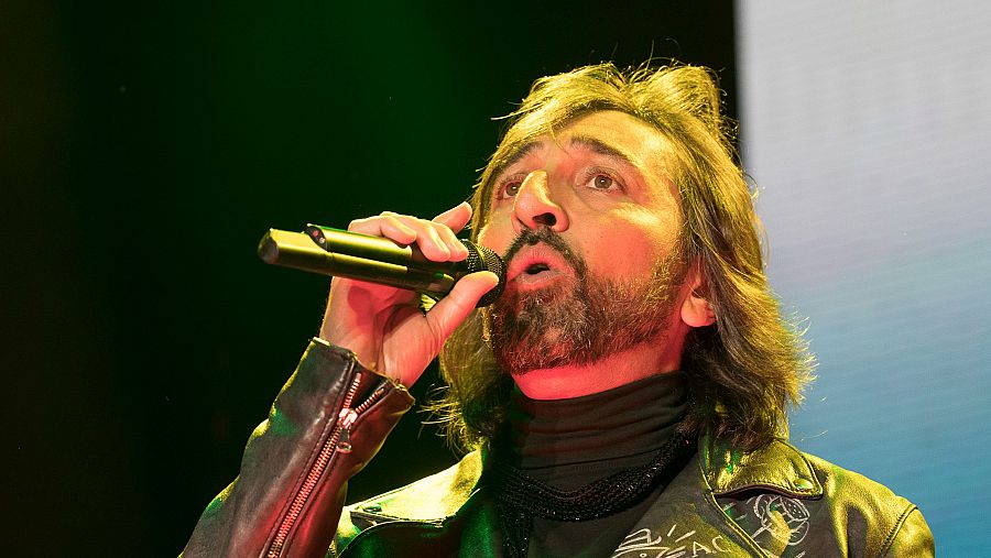 Antonio Carmona de Ketama canta al micrófono, con chaqueta negra y pelo largo castaño, sobre fondo oscuro con detalles azulados.