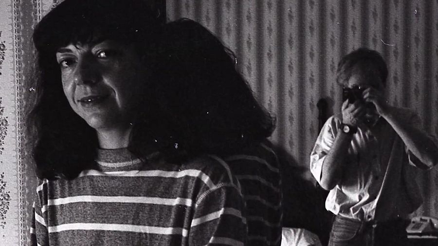 Retrato en blanco y negro de una mujer con jersey a rayas y sombrero, sonriendo ligeramente mientras es fotografiada por un hombre con camisa blanca.