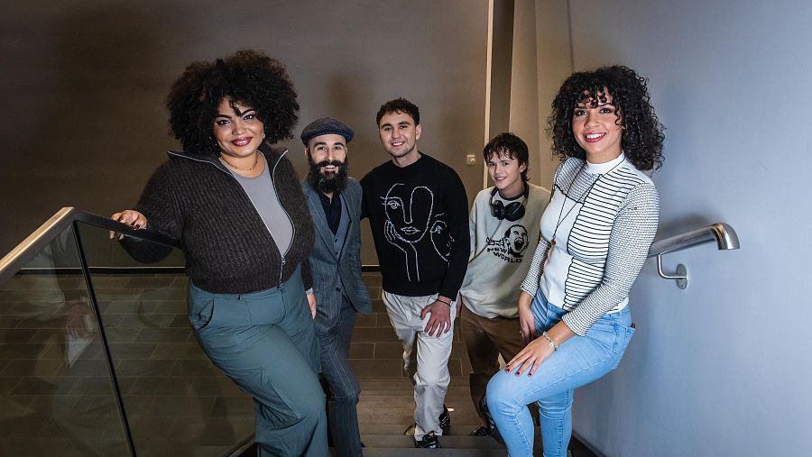 Grupo musical posando en escalera interior; cinco integrantes con ropa informal, estilos variados.  Ambiente relajado.