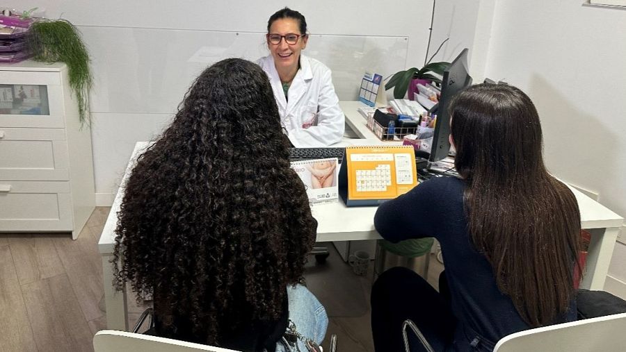 Ginecóloga en consulta, sonriendo a dos pacientes con el pelo largo y oscuro.  Sobre el escritorio se observa un ordenador y un calendario.