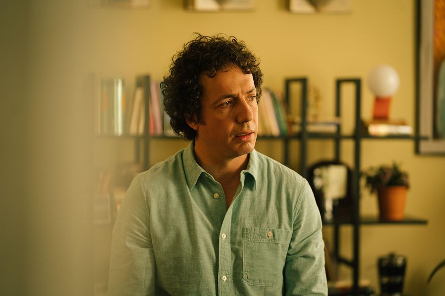 Primer plano de Vito Sanz interpretando a Pablo, con expresión seria y camisa verde. Fondo con estantería y objetos decorativos. Ambiente cálido y tenso.