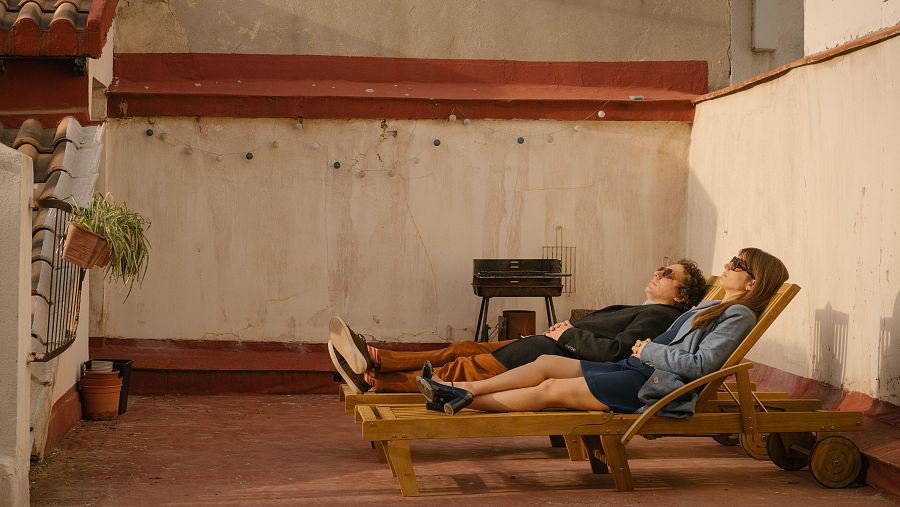 Dos personas relajadas en tumbonas de madera en una terraza con barbacoa, plantas y una pared con guirnalda. Él lleva pantalones mostaza y chaqueta oscura; ella, vestido azul y chaqueta vaquera. Ambos usan gafas de sol.