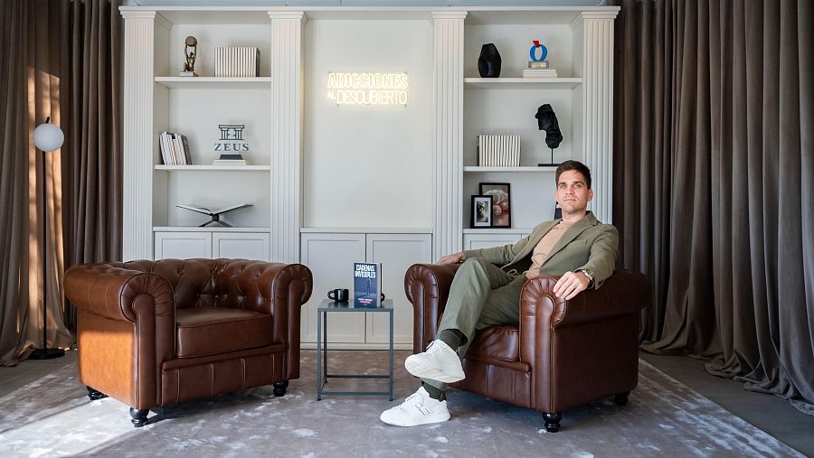 Ex adicto y terapeuta en su consulta, con sillones, estanterías con libros sobre adicciones y un letrero de neón que dice 