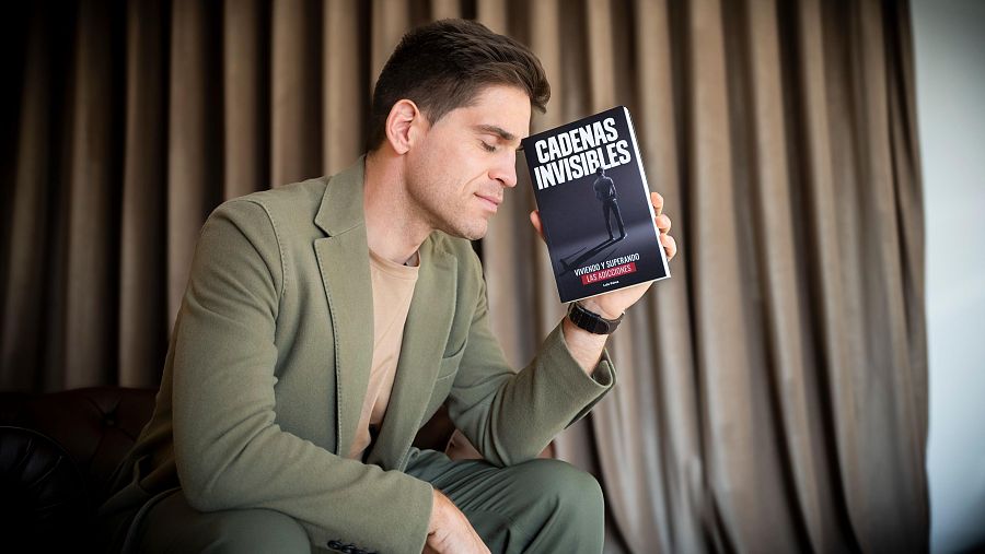 Autor sentado en sillón, traje verde oliva, sosteniendo su libro con portada negra y título 