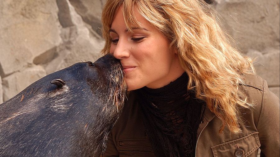 Cantante con cabello rubio y largo besa a una foca en un entorno rocoso. Lleva chaqueta marrón oliva sobre ropa negra.