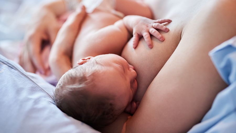 Lactancia materna: bebé de piel clara y cabello rubio mamando plácidamente del pecho de su madre, ambos sobre una superficie azul clara.
