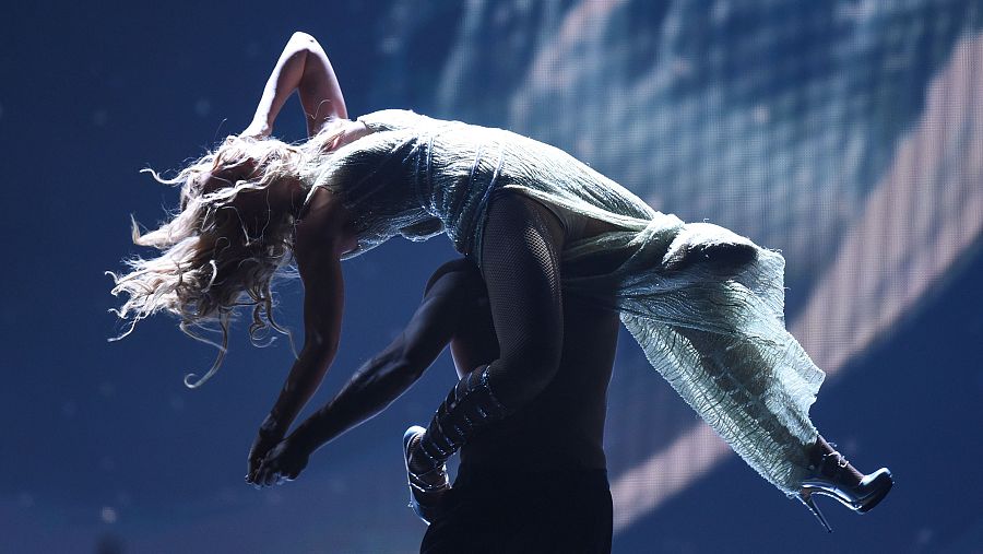 Cantante con vestido largo sostenida por un bailarín, cuerpo arqueado, melena larga, fondo oscuro.  Momento de gran intensidad en una actuación.