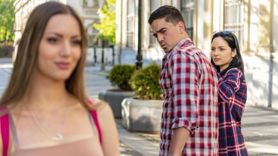 Escena callejera con tres jóvenes: un hombre con camisa a cuadros y dos mujeres. Él camina con una de ellas, pero mira a la otra, recreando el meme del 