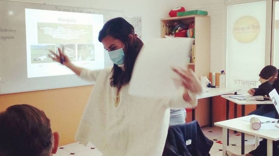 Auxiliar de conversación imparte clase de inglés sobre verduras, con presentación sobre Mongolia proyectada en pizarra.  Se observa un logo 'Trinity' en la ventana.