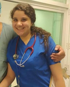 Joven con uniforme médico azul y estetoscopio, sonriendo en un pasillo hospitalario.  Una mano descansa sobre su hombro; se ve un sujetapapeles.