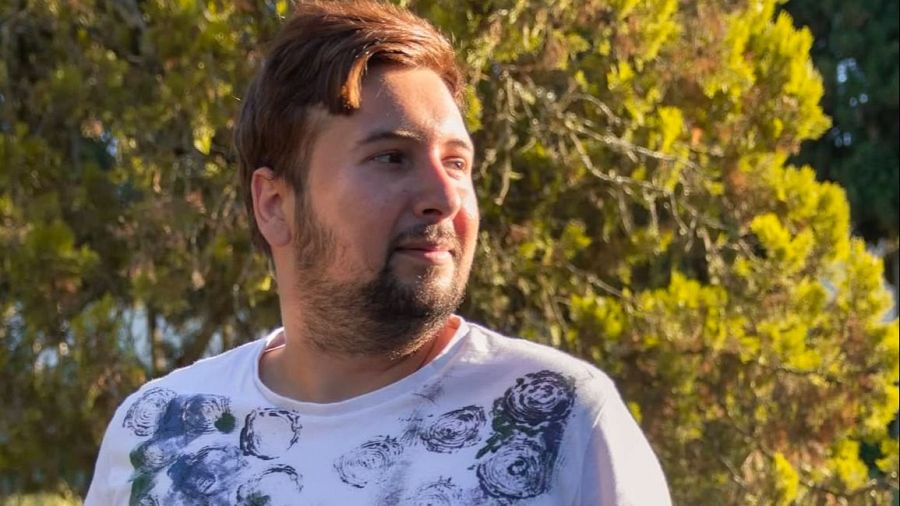 Retrato al aire libre de un hombre con pelo castaño rojizo, barba y camiseta blanca estampada, junto a texto que menciona un máster de reporterismo.