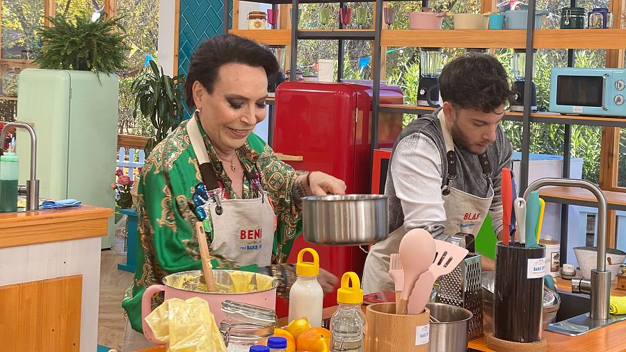 Dos personas con delantales, una mujer con blusa verde y un hombre con camiseta gris, cocinan en un set de cocina luminoso con electrodomésticos pastel.