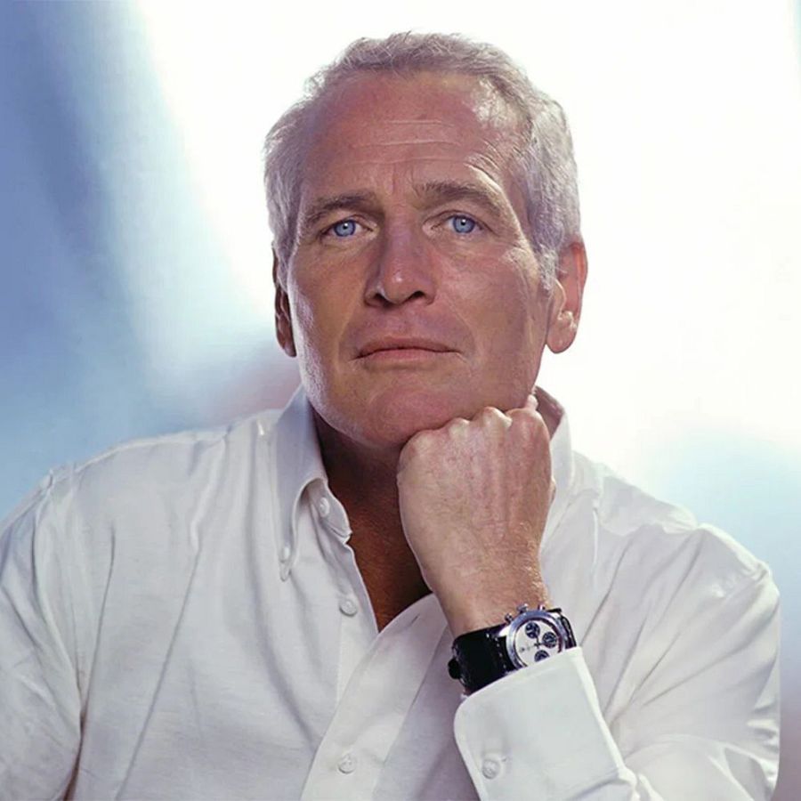 Retrato en primer plano de Paul Newman, hombre mayor con cabello gris, ojos azules, camisa blanca y reloj. Expresión seria y reflexiva.