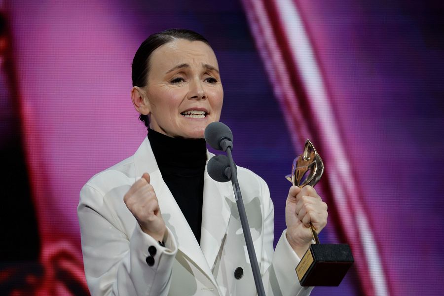 Actriz con chaqueta blanca y jersey negro, sosteniendo un premio con una base oscura y figura plateada, mostrando alegría y dando un discurso.