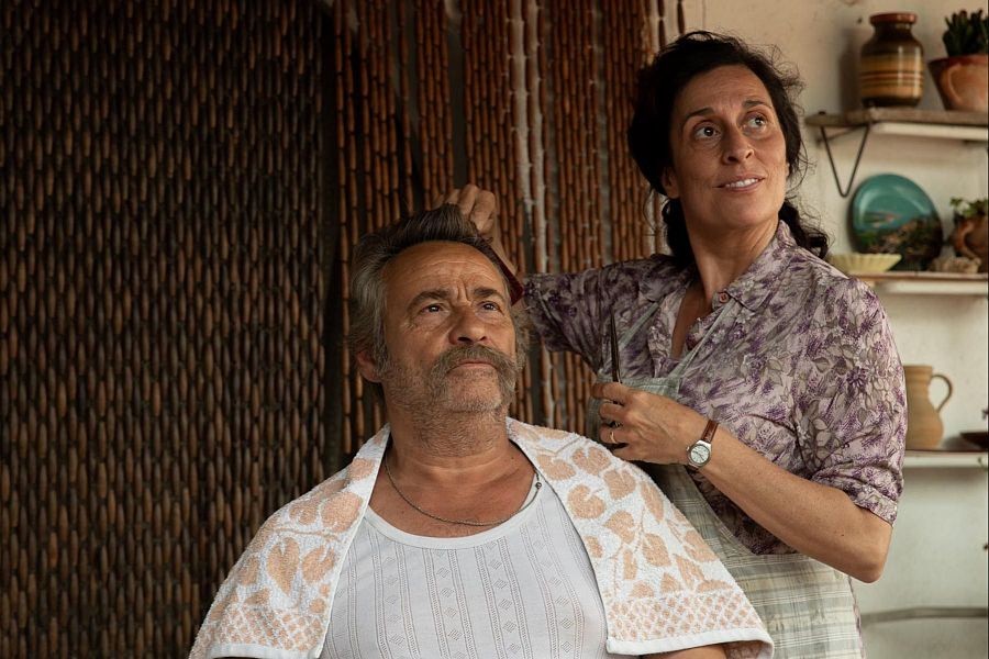 Escena de 'El 47' con Eduard Fernández sentado, con camiseta blanca y toalla, recibiendo un corte de pelo de Clara Segura, quien lleva camisa estampada y delantal. Ambiente hogareño.